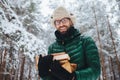 Handsome bearded male in eyewear and warm hat with anorak, holds firewood, poses against trees covered with white sparkling snow,