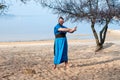 Man with bun on head in blue kimono with sword standing on sand and looking at camera