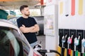 Handsome bearded man cross the arms during refueling car. Male looking on the scoreboard at gas station