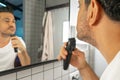 Handsome bearded man is shaving his face with trimmer machine in front of bathroom mirror Royalty Free Stock Photo