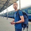 Handsome bearded man tourist with backpack stand on railway station platform and waiting for train. Travel concept. Square