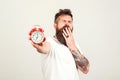 Handsome bearded man yawning over white wall. It`s 7 oclock in the morning. Hipster wears white t-shirt, holding alarm clock at