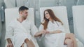 Handsome bearded man and his girlfriend wearing bathrobes are sitting in chairs in relaxation room in day spa, drinking Royalty Free Stock Photo