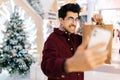 Handsome bearded young man looking at camera taking selfie on phone posing with paper bags with gifts standing in hall Royalty Free Stock Photo