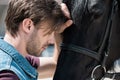 Handsome bearded young man touching purebred brown horse outdoors