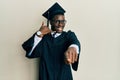 Handsome black man wearing graduation cap and ceremony robe smiling doing talking on the telephone gesture and pointing to you Royalty Free Stock Photo