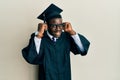 Handsome black man wearing graduation cap and ceremony robe smiling pulling ears with fingers, funny gesture Royalty Free Stock Photo