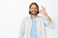 Handsome blond man with beard showing okay sign, standing in white shirt against studio background Royalty Free Stock Photo