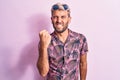 Handsome blond man on vacation wearing casual shirt and sunglasses over pink background angry and mad raising fist frustrated and Royalty Free Stock Photo