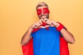 Handsome blond man wearing super hero costume with mask and cape over yellow background smiling in love doing heart symbol shape Royalty Free Stock Photo