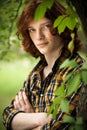 Handsome blue-eyed red-haired man leaning against tree trunk with arms crossed. Young man in plaid shirt stands among Royalty Free Stock Photo