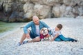 Handsome bold stylish dressed in jeans man in blue cap sitting on a beach sea side together with cute son boy teen andpretty engli