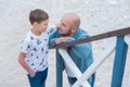 Handsome bold stylish dressed in jeans man in blue cap sitting on a beach sea side together with cute son boy teen andpretty engli