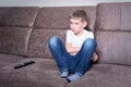 A handsome boy sits on a sofa with a remote control and watches TV. The concept of children`s leisure time