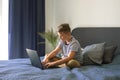 Handsome boy using his laptop for doing homework, sitting on his bed in modern interior. Studying online during Royalty Free Stock Photo
