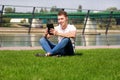 Handsome boy using tablet and headphones, drinking coffee to go, man sitting on grass and enjoying nice sunny day by river side.