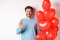 Handsome boyfriend saying hello, bring romantic red heart balloons on date, waving hand and smiling, standing over white