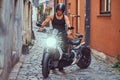 A handsome brutal biker, standing near a motorcycle, in a narrow old Europe street.