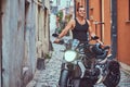 A handsome brutal biker, standing near a motorcycle, in a narrow old Europe street.