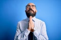 Handsome business bald man with beard wearing elegant tie and glasses over blue background begging and praying with hands together