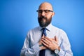 Handsome business bald man with beard wearing elegant tie and glasses over blue background smiling with hands on chest with closed