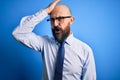 Handsome business bald man with beard wearing elegant tie and glasses over blue background surprised with hand on head for