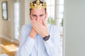 Handsome business man wearing golden crown as a king or prince shocked covering mouth with hands for mistake