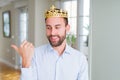 Handsome business man wearing golden crown as a king or prince smiling with happy face looking and pointing to the side with thumb