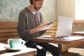 Handsome businessman with long hair wearing sweater calling by smartphone sitting in sunny cafe, using laptop having coffee alone