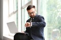 Handsome businessman looking at watch while working with laptop near window Royalty Free Stock Photo