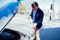 A handsome businessman wearing blue blazer lifting up the hood of his car and checking the oil level on a sunny day parked on a