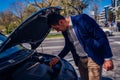 A handsome businessman wearing blue blazer lifting up the hood of his car and checking the oil level on a sunny day parked on a