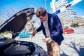 A handsome businessman wearing blue blazer lifting up the hood of his car and checking the oil level on a sunny day parked on a