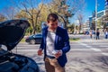 A handsome businessman wearing blue blazer lifting up the hood of his car and checking the oil level on a sunny day parked on a