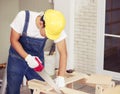 Handsome carpenter or repairman wearing yellow helmet working in wood workshop using handsaw cutting wood plank