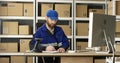 Handsome Caucasian mailman in blue iniform and cap sitting at table in postal office store and working at computer