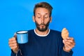 Handsome caucasian man with beard drinking a cup of coffee and croissant making fish face with mouth and squinting eyes, crazy and