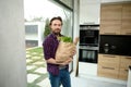 Handsome Caucasian man, charming guy, stands in a home kitchen with a cardboard bag full of healthy food and whole grain bread, Royalty Free Stock Photo