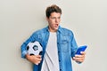 Handsome caucasian man holding football ball looking at smartphone afraid and shocked with surprise and amazed expression, fear Royalty Free Stock Photo