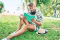 Handsome caucasian man wearing summer hat and flowers shirt smiling happy sitting on the grass reading a book Royalty Free Stock Photo