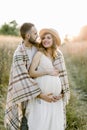 Handsome Caucasian man hugging his attractive pregnant woman in straw hat and white dress in sunny day in field. Happy Royalty Free Stock Photo