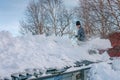 Handsome Caucasian teenager with shovel remove snow from roof of totally snow covered house, cabin in mountains. Cloudy warm day,