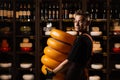 Handsome cheese sommelier with yellow wheels of cheese in shop. Worker of food cheese store carrying rounds of cheese.