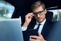 Handsome confident man in full suit looking at his smart phone while sitting in the car and using laptop. Royalty Free Stock Photo