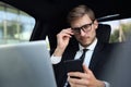 Handsome confident man in full suit looking at his smart phone while sitting in the car and using laptop Royalty Free Stock Photo