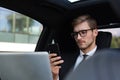 Handsome confident man in full suit looking at his smart phone while sitting in the car and using laptop Royalty Free Stock Photo