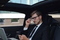 Handsome confident man in full suit looking at his smart phone while sitting in the car and using laptop Royalty Free Stock Photo