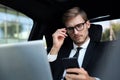 Handsome confident man in full suit looking at his smart phone while sitting in the car and using laptop Royalty Free Stock Photo