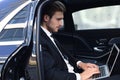 Handsome confident man in full suit looking at his smart phone while sitting in the car and using laptop Royalty Free Stock Photo