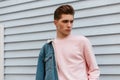 Handsome cute young man model in trendy pink sweatshirt in fashion denim blue youth jacket posing near white wall outdoors. Urban Royalty Free Stock Photo